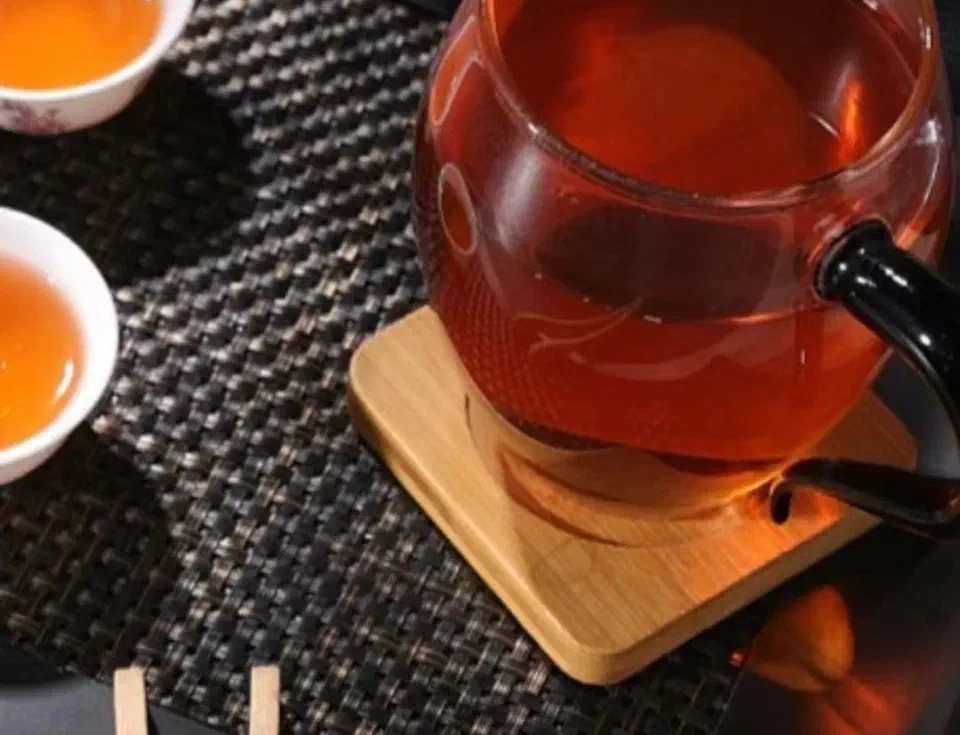 Reddish-amber brewed dark tea in a glass pitcher on a bamboo coaster, beside two white cups of tea.